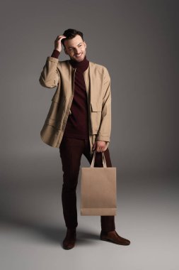Fashionable man in brown jacket holding shopping bag on grey background