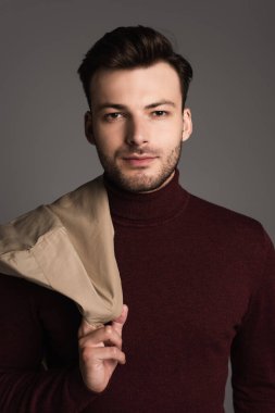 Portrait of brunette man in turtleneck holding jacket isolated on grey 