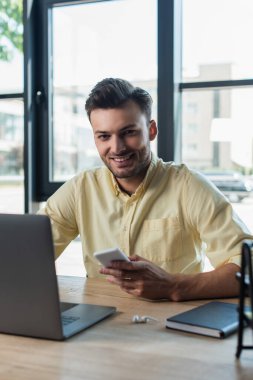 Smiling businessman looking at camera while holding smartphone near laptop in office 