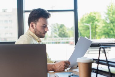 Side view of businessman holding paper folder near blurred laptop and coffee to go in office 