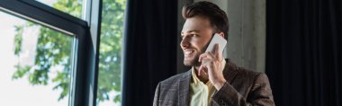 Cheerful manager talking on mobile phone near window in office, banner 