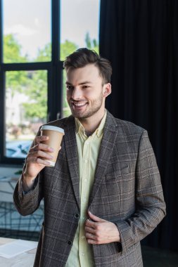 Cheerful businessman in jacket holding takeaway drink in office 