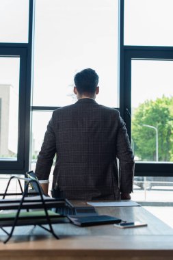Back view of businessman in jacket standing near devices and papers on working table in office 