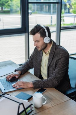 Businessman in headphones using laptop near papers and cup on table in office 