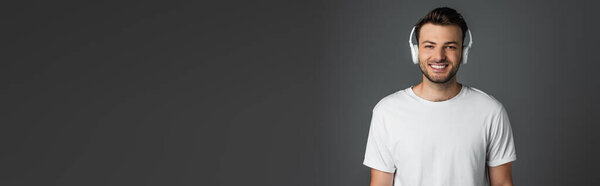 Positive man in white headphones looking at camera isolated on grey, banner 