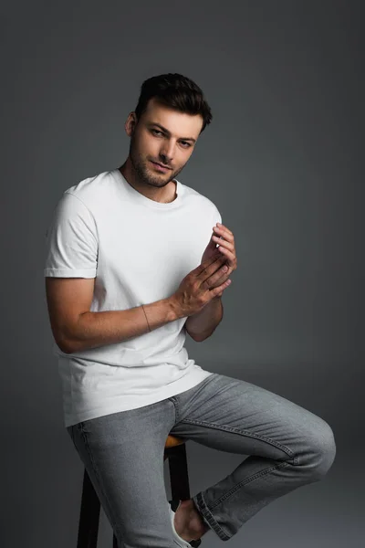 Brunette man in jeans and t-shirt posing while sitting on chair isolated on grey