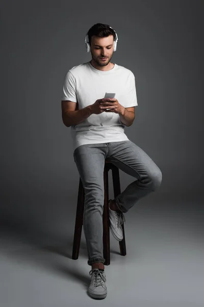 Full length of young man in jeans and t-shirt using smartphone and headphones while sitting on chair on grey background