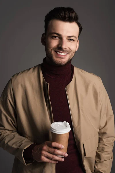 stock image Smiling brunette man in autumn outfit holding paper cup and looking at camera isolated on grey 