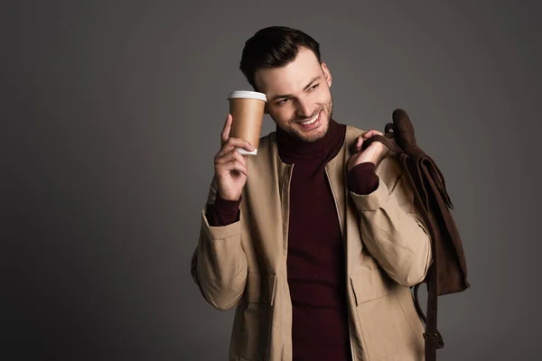 Positive man in autumn outfit holding backpack and paper cup isolated on grey