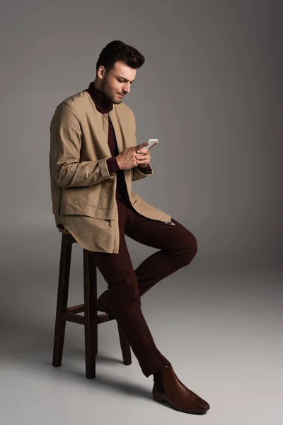 Young man in autumn outfit using smartphone while sitting on chair on grey background