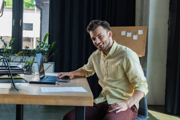 Χαμογελώντας Επιχειρηματίας Κοιτάζοντας Κάμερα Ενώ Χρησιμοποιώντας Φορητό Υπολογιστή Στο Γραφείο — Φωτογραφία Αρχείου