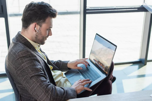 Side view of businessman in earphone using laptop in office