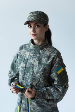 Soldier in cap and uniform holding blue and yellow ribbon isolated on grey 