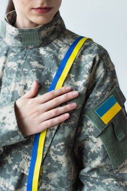 Cropped view of soldier with ukrainian flag on chevron touching blue and yellow ribbon isolated on grey 