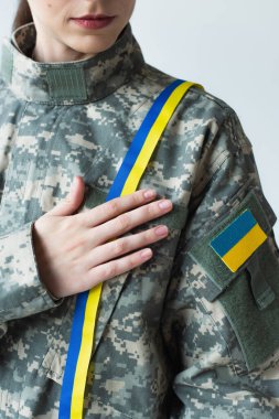 Cropped view of soldier with ukrainian flag on chevron touching blue and yellow ribbon isolated on grey  clipart