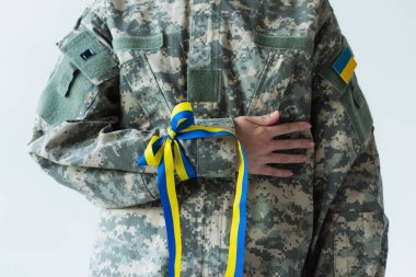 Cropped view of soldier with ukrainian flag and blue and yellow ribbon on hand isolated on grey 