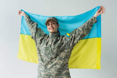 Smiling soldier in uniform holding ukrainian flag isolated on grey  clipart