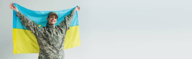 Happy servicewoman in camouflage uniform holding ukrainian flag isolated on grey, banner 