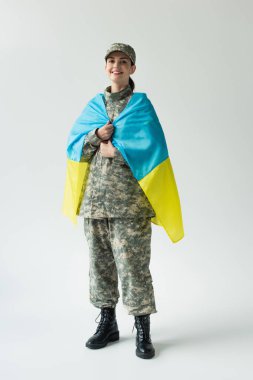 Happy soldier covering with ukrainian flag on grey background clipart