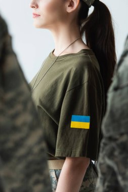 Cropped view of servicewoman with ukrainian flag on chevron standing near blurred soldiers isolated on grey 