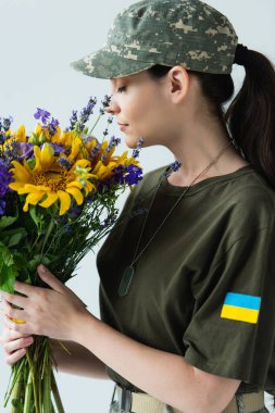 Side view of servicewoman with ukrainian flag on chevron smelling flowers isolated on grey 