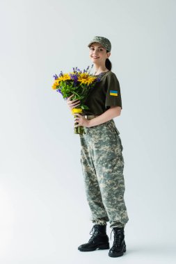 Positive soldier in uniform with ukrainian flag holding bouquet on grey background clipart