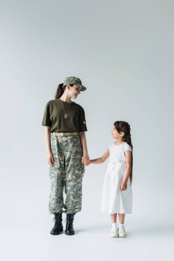 Side view of soldier with ukrainian flag on uniform holding hand of daughter on grey background