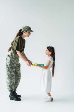 Side view of kid holding ukrainian flag near mother in military uniform on grey background