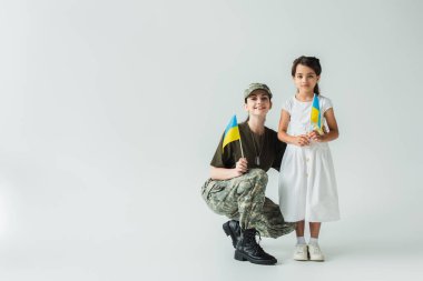 Soldier in uniform and daughter holding ukrainian flags on grey background