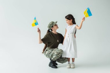 Side view of smiling parent in military uniform and child holding ukrainian flags on grey background