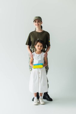 Smiling woman in camouflage uniform hugging daughter with ukrainian flag on grey background