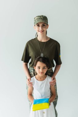 Kid holding ukrainian flag near smiling parent in camouflage uniform isolated on grey 