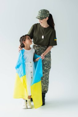 Smiling mother in camouflage uniform holding ukrainian flag and hugging daughter on grey background