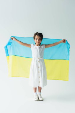 Kid holding ukrainian flag and looking at camera on grey background clipart