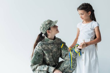 Kid holding blue and yellow ribbon and looking at mom in military uniform isolated on grey 