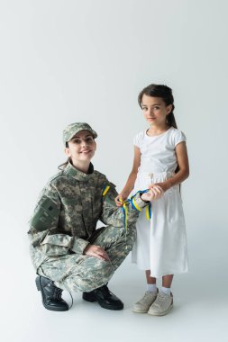 Smiling child holding blue and yellow ribbon near mom in camouflage uniform on grey background