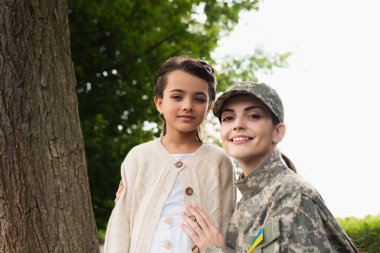 Smiling soldier with ukrainian flag on chevron hugging daughter outdoors 