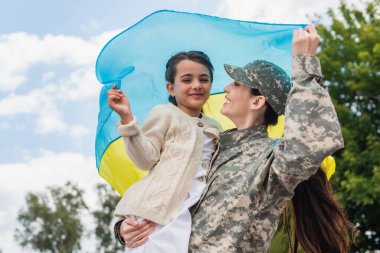 cheerful military woman with daughter holding ukrainian flag outdoors