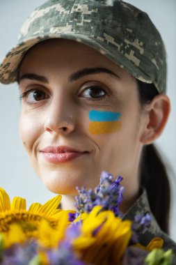 close up portrait of happy ukrainian servicewoman near blue and yellow flowers isolated on grey