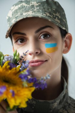 portrait of positive military woman looking at camera near flowers isolated on grey