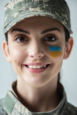 close up portrait of military woman with ukrainian flag on face smiling at camera isolated on grey