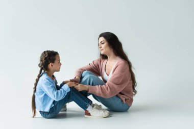 side view of mom and daughter in jeans holding hands while sitting on grey background