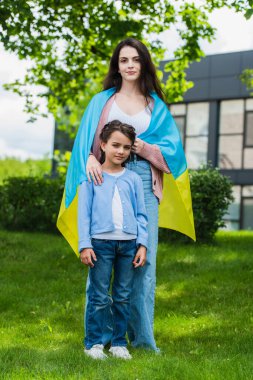 woman covered with ukrainian flag looking at camera near daughter in park