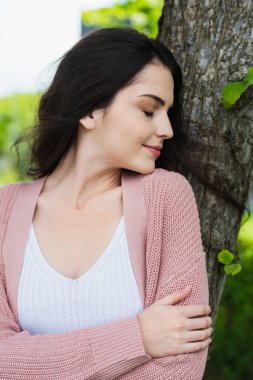 happy brunette woman with closed eyes standing near tree trunk 