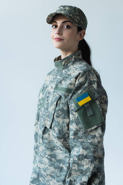 Smiling servicewoman in camouflage uniform with ukrainian flag looking at camera isolated on grey 