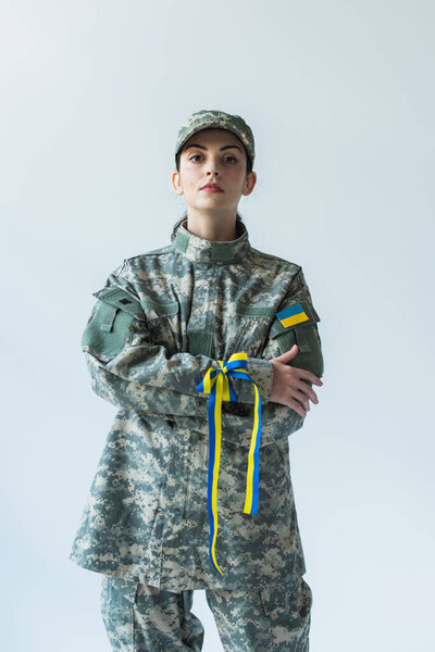 Soldier in uniform with ukrainian flag and blue and yellow ribbon crossing arms isolated on grey 
