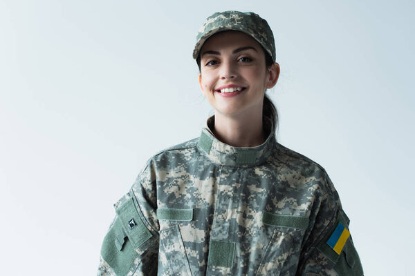 Cheerful soldier in uniform with ukrainian flag looking at camera isolated on grey 