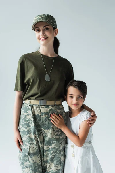 Smiling servicewoman in military uniform hugging child isolated on grey