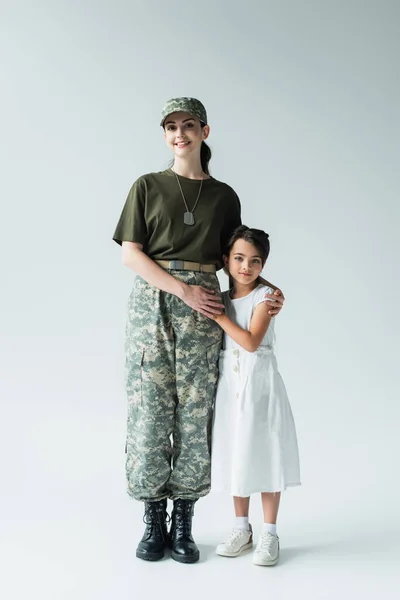 Daughter hugging smiling mother in military uniform on grey background