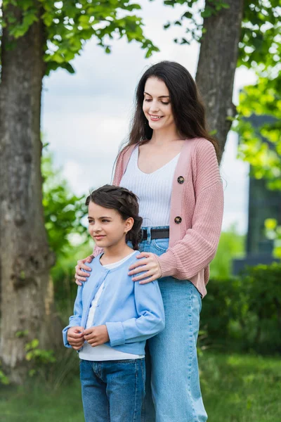 smiling woman embracing shoulders of pensive daughter in park